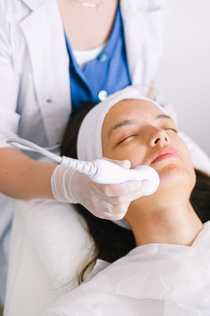 About Professional female cosmetologist wearing medical uniform doing ultrasonic face cleaning for relaxed young female customer in modern beauty clinic